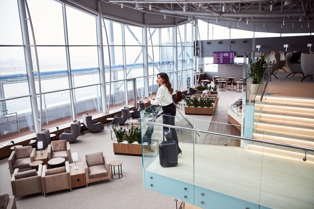A traveler stands in an upscale airport lounge, gazing out over the tarmac with her suitcase nearby.