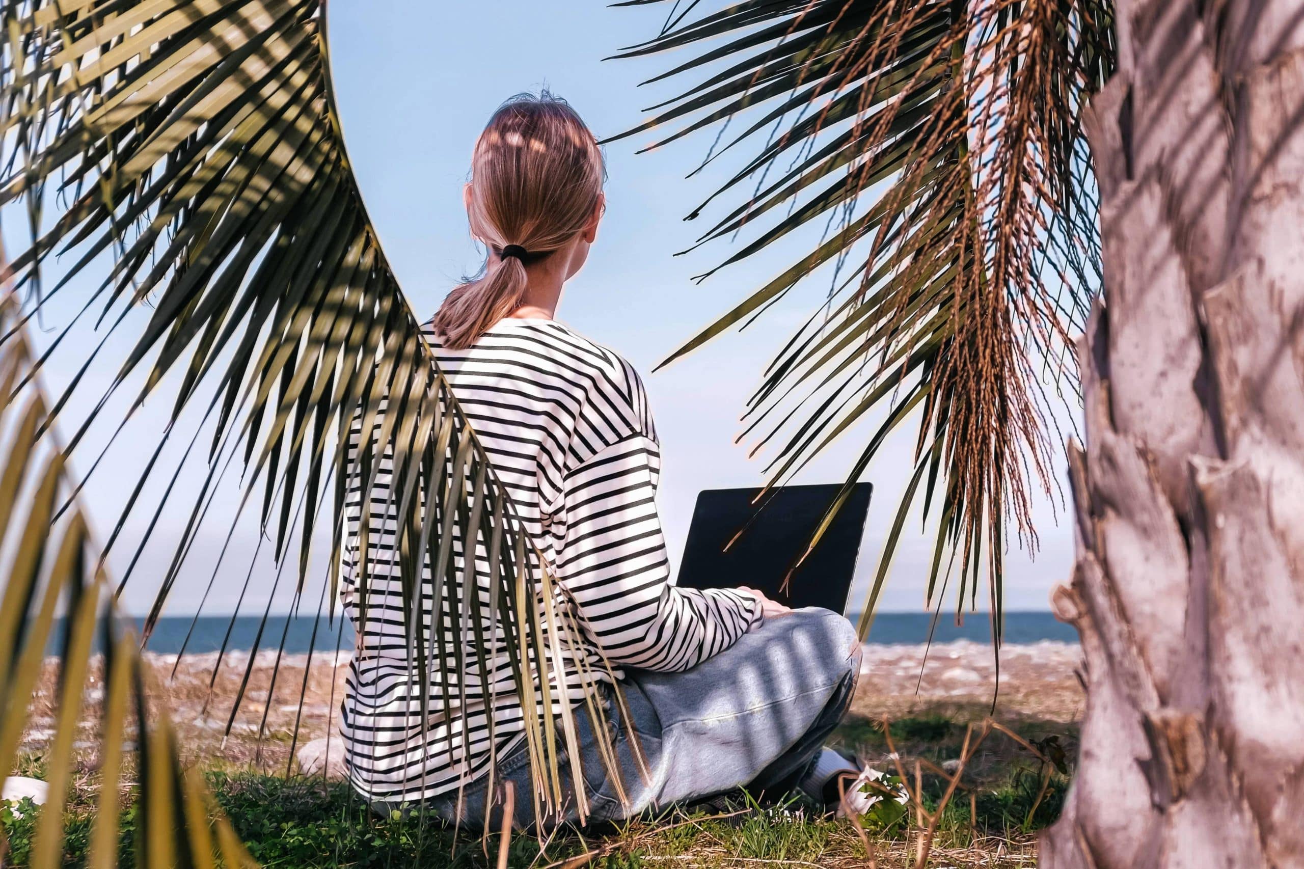 A woman sitting on the grass near the ocean working on a laptop